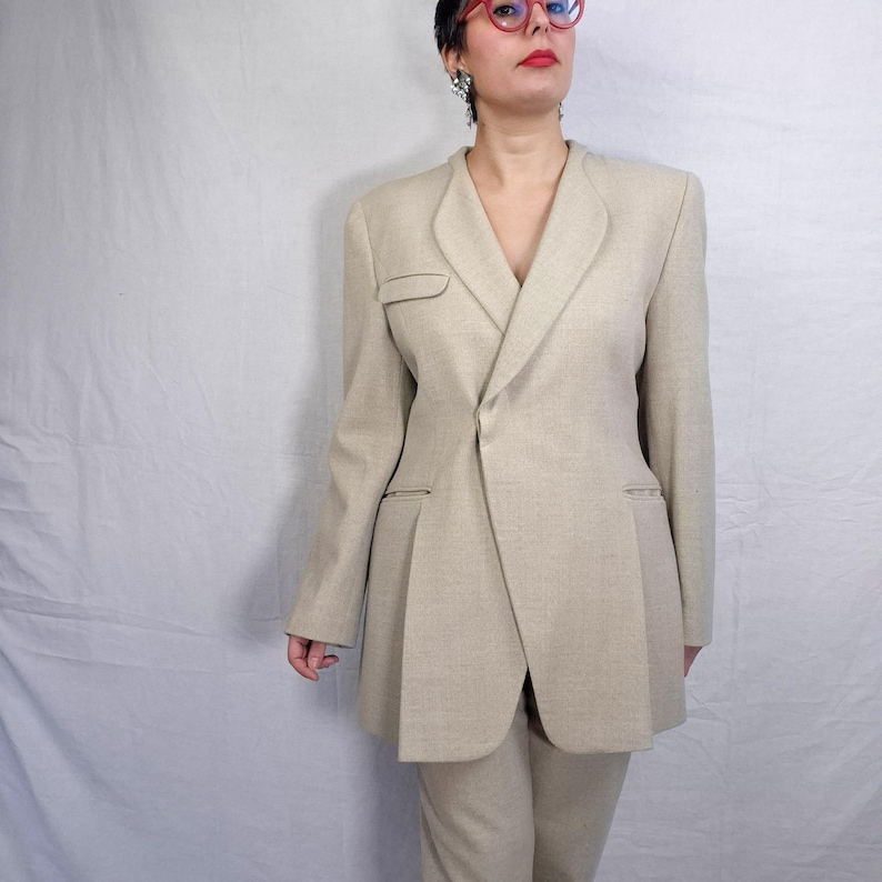Puede incluir: Un traje de dos piezas beige. La chaqueta a medida presenta un cierre asim&eacute;trico y solapas con muescas. La chaqueta tambi&eacute;n tiene un peque&ntilde;o bolsillo y un dise&ntilde;o plisado. El traje incluye pantalones a juego. El traje est&aacute; hecho de una tela texturizada.