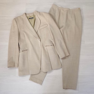 Puede incluir: Un traje de dos piezas beige, compuesto por una americana y un pantal&oacute;n a juego. La americana tiene solapas con muescas, dos bolsillos delanteros y un cierre de un solo bot&oacute;n. El pantal&oacute;n es de corte recto. El traje est&aacute; hecho de una tela texturizada.