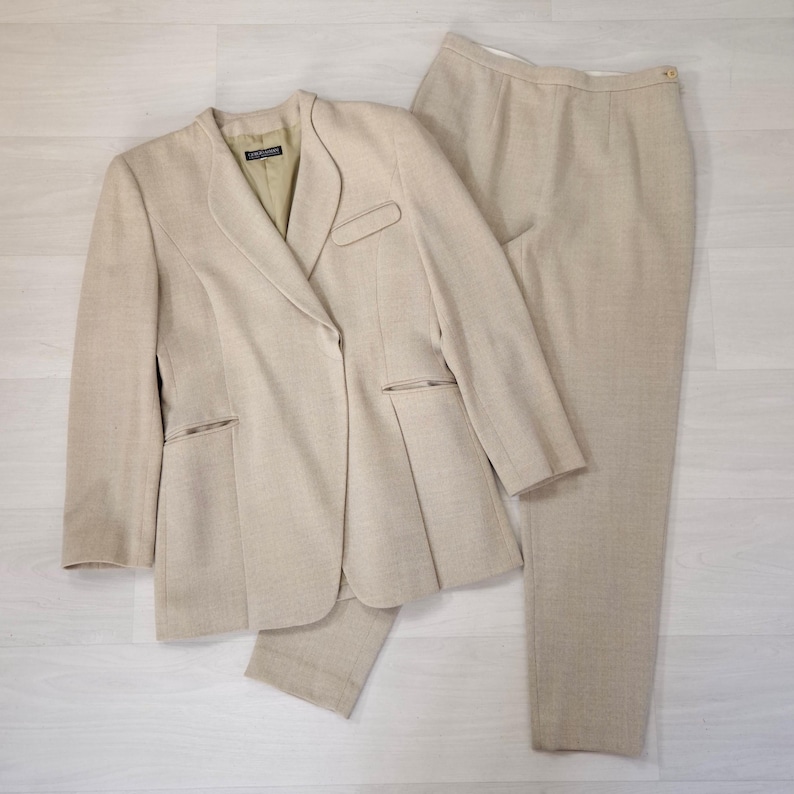 Puede incluir: Un traje de dos piezas beige, compuesto por una americana y un pantal&oacute;n a juego. La americana tiene solapas con muescas, dos bolsillos delanteros y un cierre de un solo bot&oacute;n. El pantal&oacute;n es de corte recto. El traje est&aacute; hecho de una tela texturizada.