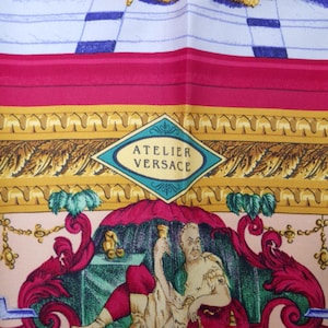 Pu&ograve; includere: Una sciarpa di seta con un design dettagliato. La sciarpa presenta un'immagine centrale di una persona in un'ambientazione classica, circondata da bordi ornati e il testo "ATELIER VERSACE". La tavolozza dei colori include rosa, oro, rosso e blu.