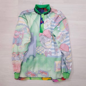 May include: A long-sleeved shirt with a colorful, cartoonish design. The shirt features a green collar and cuffs, with a button-up placket. The pattern includes various figures and shapes in shades of green, blue, pink, and yellow.