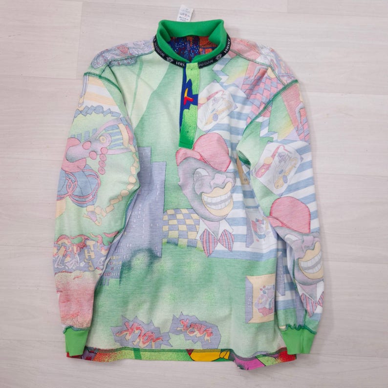 May include: A long-sleeved shirt with a colorful, cartoonish design. The shirt features a green collar and cuffs, with a button-up placket. The pattern includes various figures and shapes in shades of green, blue, pink, and yellow.