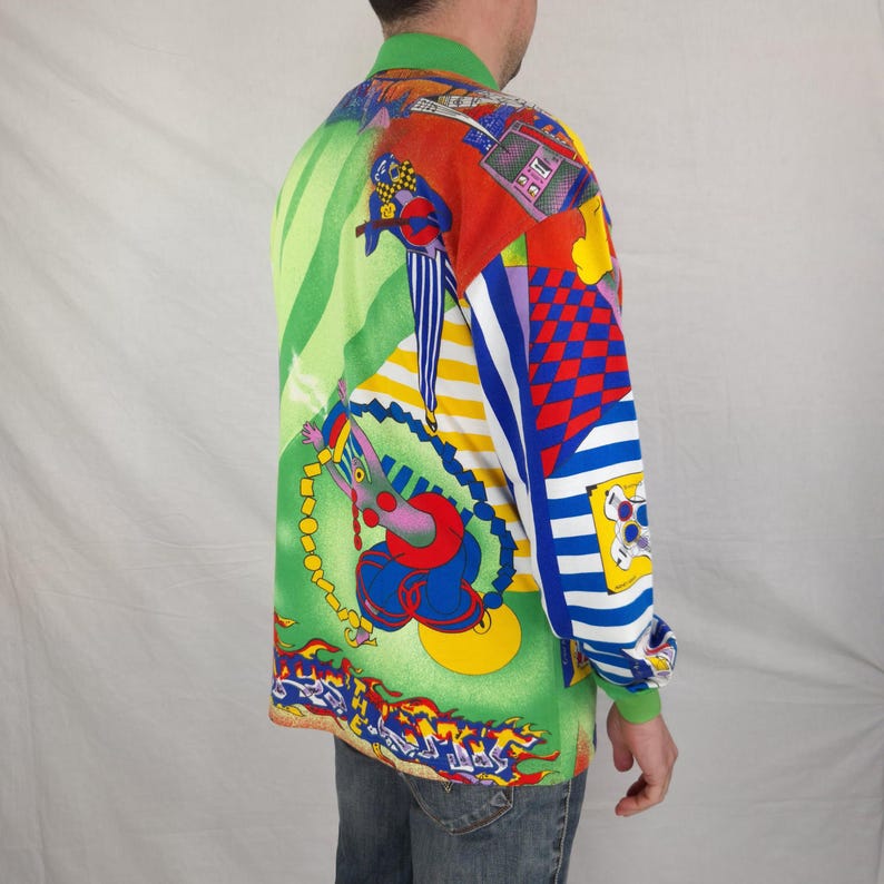 May include: A long-sleeved shirt with a vibrant, colorful design. The shirt features a green collar and cuffs, with a patchwork of patterns including stripes, checkerboards, and abstract figures in red, blue, yellow, and green. The shirt is worn by a person.
