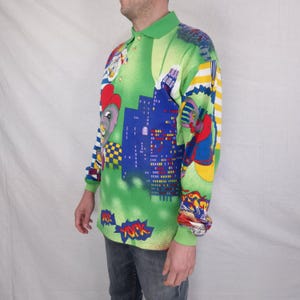 May include: A long-sleeved polo shirt with a vibrant green base, featuring a cityscape and cartoon character design. The shirt has a green collar and cuffs, with a button-up neckline. The design includes blue buildings, cartoon figures, and the word "YORK".