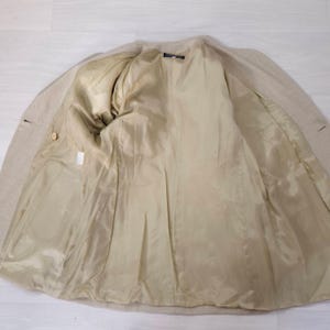 Puede incluir: Blazer beige con forro interior de color beige claro. El interior de la chaqueta es visible, mostrando el forro completo y la confecci&oacute;n a medida. El blazer tiene dos botones y un dise&ntilde;o de solapa cl&aacute;sico.