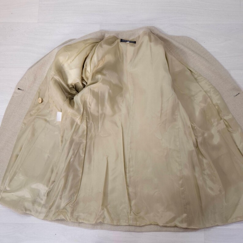 Puede incluir: Blazer beige con forro interior de color beige claro. El interior de la chaqueta es visible, mostrando el forro completo y la confecci&oacute;n a medida. El blazer tiene dos botones y un dise&ntilde;o de solapa cl&aacute;sico.