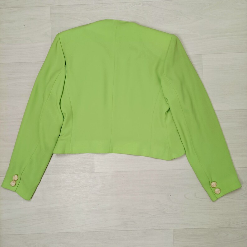 May include: A bright green blazer with long sleeves and gold buttons. The blazer is cropped and has a simple design.