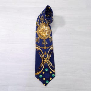 Pode incluir: Uma gravata azul marinho com um design dourado e branco ornamentado. A gravata apresenta um grande emblema circular e elementos decorativos menores. A parte inferior da gravata tem um padrão de círculos azuis e verdes.
