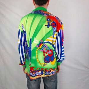 May include: A long-sleeved shirt with a vibrant, colorful design. The shirt features a green collar and cuffs, with a bold graphic on the back. The graphic includes a cartoon figure, cityscapes, and stripes in shades of green, red, blue, and yellow.