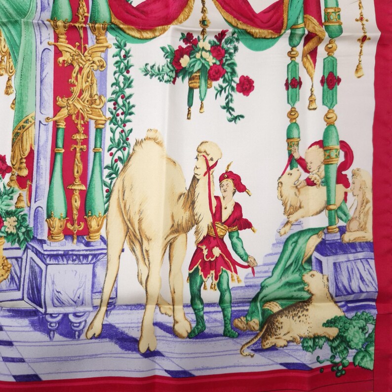 Pu&ograve; includere: Una sciarpa di seta bianca con un'illustrazione colorata di un cammello, figure in abiti rossi e verdi ed elementi architettonici. La sciarpa ha un bordo rosso e dettagli decorativi floreali e di tendaggi.