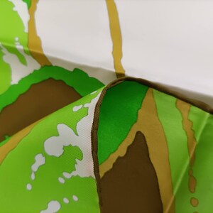 Op de afbeelding: Close-up van een groene, bruine en witte stof met een bruine naad. De stof heeft een zachte, vloeiende textuur.