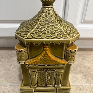 May include: A green ceramic piggy bank shaped like a small castle with a brown roof and a door. The castle has a small tower on each side.