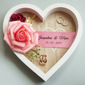 Könnte beinhalten: Weiße herzförmige Box mit einer rosa Rose, einer burgunderfarbenen Rose, einem goldenen Blatt und zwei goldenen Ringen. Die Box hat ein rosa Band mit dem Text "Jacqueline & Marc 21.01.2021".