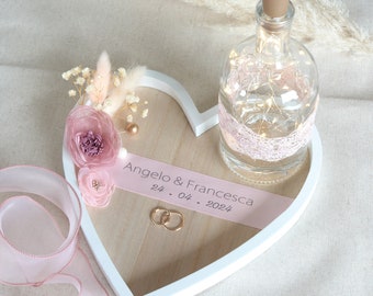 May include: A white heart-shaped tray with a wooden base, decorated with pink flowers, gold beads, and a pink ribbon with the text "Angelo & Francesca 24. 04. 2024". A clear glass bottle with a cork top and a pink lace ribbon is on the tray. Two gold rings are on the tray.