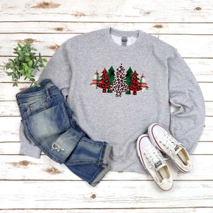 May include: A gray sweatshirt with a Christmas tree design featuring a red and green plaid pattern, leopard print, and green and red plaid. The sweatshirt is paired with blue jeans and white sneakers.