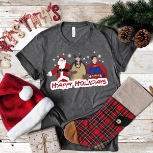 May include: A dark gray t-shirt with a Christmas-themed design featuring Santa Claus, a donkey, and Superman. The text "Happy Holidays" is printed below the characters. The shirt is surrounded by Christmas decorations, including a Santa hat, a stocking, and a wooden sign that says "Christmas".