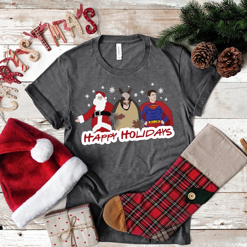 May include: A dark gray t-shirt with a Christmas-themed design featuring Santa Claus, a donkey, and Superman. The text "Happy Holidays" is printed below the characters. The shirt is surrounded by Christmas decorations, including a Santa hat, a stocking, and a wooden sign that says "Christmas".