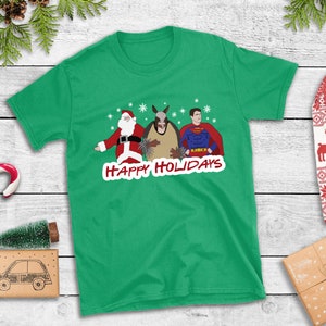 May include: Green t-shirt with a Christmas-themed design featuring Santa Claus, a man dressed as Superman, and a man in an armadillo costume. The text "Happy Holidays" is printed in red on the shirt.