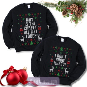 May include: Two black sweatshirts with Christmas-themed designs. One says "WHY IS THE CARPET ALL WET TODD?" and the other says "I DON'T KNOW MARGO!". Both have red, green, and white snowflake, gift, tree, and reindeer patterns. Festive holiday apparel.