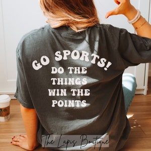May include: A charcoal grey t-shirt with white text that reads "GO SPORTS! DO THE THINGS WIN THE POINTS". The text is in a retro font style.