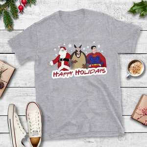 May include: A gray t-shirt with a Christmas-themed design featuring Santa Claus, a person wearing a donkey head, and Superman. The text "Happy Holidays" is printed in red below the characters.