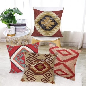 May include: A collection of decorative throw pillows with geometric patterns. The pillows feature a variety of colors, including red, beige, and teal. The designs include diamonds, squares, and Aztec-inspired motifs. The pillows are arranged on a white rug and a wooden stool.