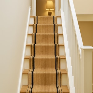 以下が含まれることがあります： 天然繊維のランナーが付いた階段。ランナーはタン色で、中央に2本の黒いストライプが入っています。階段には白い蹴上げと木製の踏み板があります。白い手すりが階段の右側にあります。階段の上にはランプがあります。