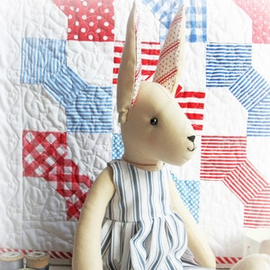 May include: A handmade doll with long ears, wearing a blue and white striped dress. The doll is sitting on a white quilt with red and blue squares.