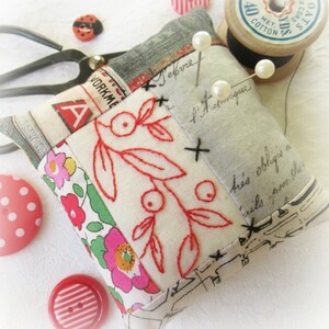 May include: A white fabric pincushion with red embroidered flowers and leaves. The pincushion is made of patchwork fabric with different patterns and colours, including a red and white floral pattern, a grey and white patterned fabric, and a white fabric with black text. The pincushion has three white pearl-like pins sticking out of it.