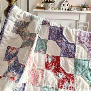 May include: A patchwork quilt with a floral pattern in shades of pink, red, blue, and purple. The quilt features a white background and is draped over a bed frame. The quilt's design includes a variety of floral prints.