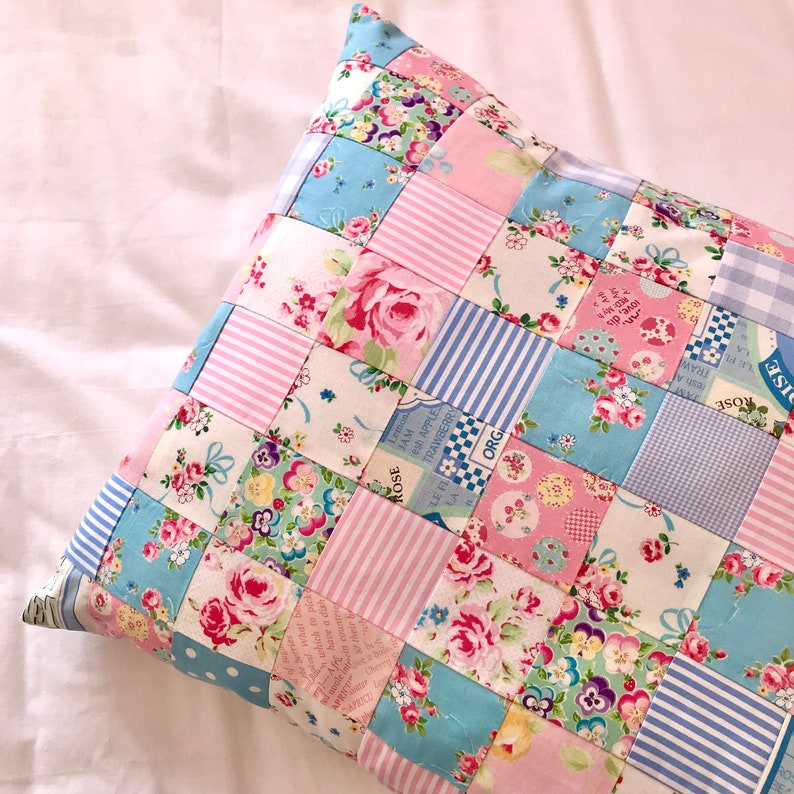 May include: A patchwork pillow with a blue, pink, and white floral pattern. The pillow is made of fabric squares sewn together. The fabric has a variety of patterns, including polka dots, stripes, and floral prints. The pillow is decorated with the words "ROSE", "TRAWBERRY", "ORG", "JAM", "Lemon", "which to pick", "love, dis", and "LE PL".