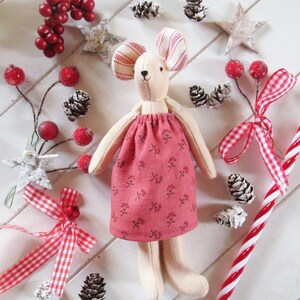 May include: A handmade plush mouse doll wearing a pink dress with a floral pattern. The doll is surrounded by red berries, pine cones, and white stars.