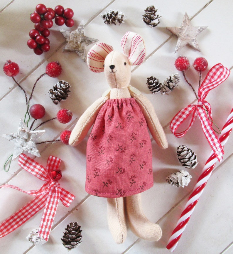 May include: A handmade plush mouse doll wearing a pink dress with a floral pattern. The doll is surrounded by red berries, pine cones, and white stars.