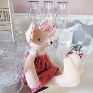 May include: A pink and white striped fabric mouse wearing a red dress with white floral print. The mouse is sitting on a white surface with clear glass jars in the background.