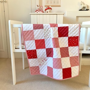 Op de afbeelding: Een patchwork quilt met een raster van afwisselende vierkanten in verschillende tinten rood en wit. De stofpatronen omvatten effen rood, rood-witte vichyruitjes, rode strepen en bloemenprints, afgewerkt met een doorgestikte ruitstructuur.
