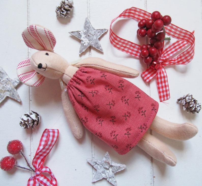May include: A handmade plush mouse toy wearing a red dress with a floral pattern. The mouse is sitting on a white surface with red and white gingham ribbon, pine cones, and silver stars.