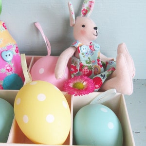 May include: A white box with a pink bunny doll sitting on top. The box contains five pastel colored Easter eggs with white polka dots.