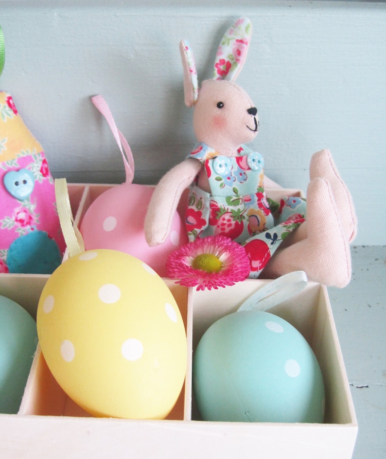 May include: A white box with a pink bunny doll sitting on top. The box contains five pastel colored Easter eggs with white polka dots.