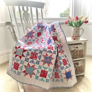 Puede incluir: Una colcha de retazos roja, blanca y azul con un patrón de estrellas está drapeada sobre una mecedora blanca. La colcha está hecha de varias telas con diferentes patrones, incluyendo lunares, rayas e estampados florales.