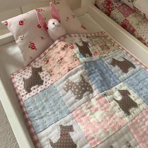 Peut inclure: Un petit lit avec un oreiller rose à motifs floraux et une courtepointe patchwork avec des appliqués de chiens. Un lapin en peluche avec des oreilles roses repose sur l'oreiller. La courtepointe présente des carrés roses, bleus et blancs.