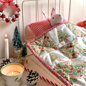 May include: A Christmas-themed display with a small white bed and a bunny plushie under a floral quilt. A red and white striped pillow supports the bunny. A small wreath, candles, and a candy cane add to the festive atmosphere.