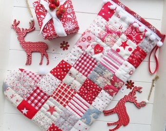 May include: A handmade Christmas stocking with a patchwork design in red and white fabrics. The stocking is decorated with stars and hearts. A red gift box, reindeer ornaments, and floral accents complete the festive scene.