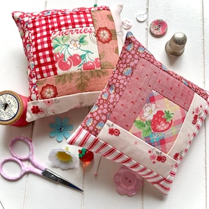 Könnte beinhalten: Zwei quadratische Nadelkissen mit Kirsch- und Erdbeermustern, zusammen mit Nähutensilien wie Schere, Knöpfen und einem Fingerhut. Die Kissen weisen rote und rosa Farbtöne mit floralen und Vichy-Mustern auf. Eine Garnrolle und dekorative Nadeln vervollständigen die Szene.