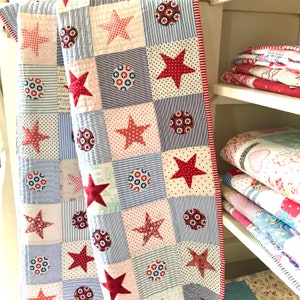 May include: A red, white and blue patchwork quilt with stars and circles. The quilt is made of different fabrics and patterns. The quilt is folded and leaning against a white shelf.