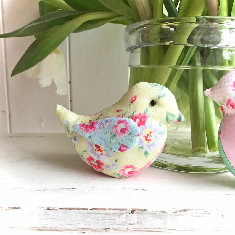 Op de afbeelding: Twee handgemaakte stoffen vogels met bloemenpatronen. De vogel links is geel met roze en blauwe bloemen. De vogel rechts is roze met witte stippen.