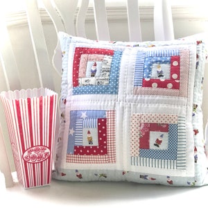 May include: A white pillow with a patchwork design featuring red, blue, and white squares with a gnome pattern. The pillow is decorated with stars, polka dots, and stripes.