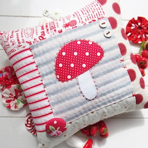 May include: A red and white striped pillow with a red and white polka dot mushroom applique. The pillow is made of fabric scraps and has a patchwork design. The pillow is sitting on a white surface.