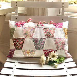 Op de afbeelding: Een wit kussen met een patchwork-ontwerp van roze, rode en groene bloemenstof. Het kussen ligt op een witte stoel met een witte lattenrug.