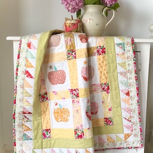 Op de afbeelding: Een groene, gele en rode patchworkdeken met een bloemenpatroon. De deken is gevouwen en gedrapeerd over een witte commode. De deken heeft een patroon van appels.