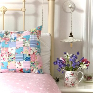May include: A pink and white polka dot bedspread with a patchwork pillow featuring floral and checkered patterns. A white ceramic lamp with a floral design hangs from the ceiling. A bouquet of purple and white flowers sits on a table next to a white pitcher.